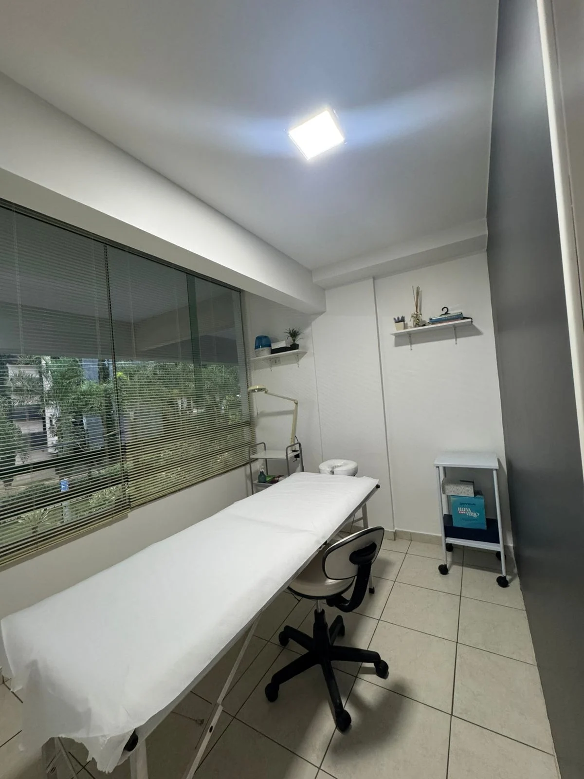 Cabine branca da LA CLINIC, com maca forrada, banqueta e janela com persiana